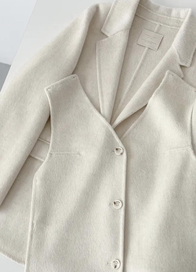 May include: Cream-colored blazer and vest set. The blazer has a classic lapel design, and the vest features a V-neck and button closure. The fabric appears soft and textured. The set is displayed on a white surface.