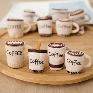 Puede incluir: Una colección de tazas de café y vasos para llevar de tela beige con detalles bordados marrones. La palabra "Coffee" está bordada en las tazas. Algunas tazas tienen asas y una tiene una pajita. Las tazas están dispuestas sobre una bandeja de madera.
