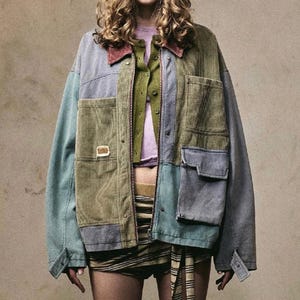 Op de afbeelding: Een veelkleurige patchworkjas in tinten blauw, groen en olijfgroen. De jas heeft een ribfluwelen textuur, meerdere zakken en een rode kraag. Het model draagt een paars shirt en een groen vest.
