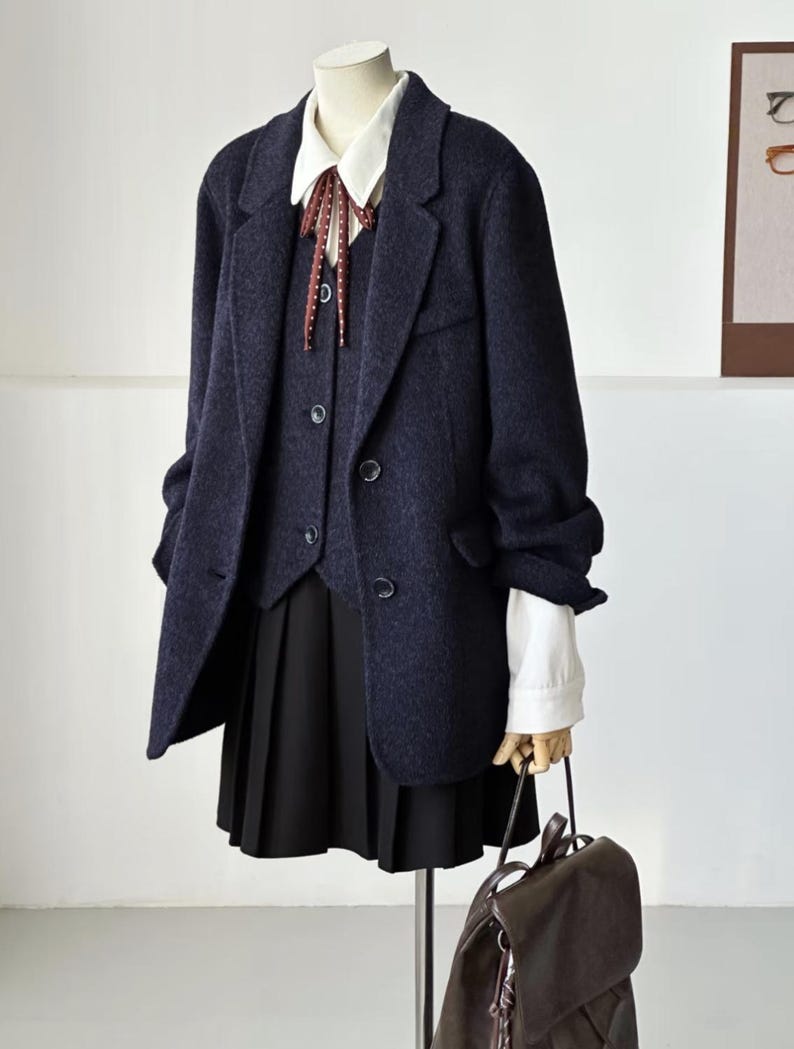May include: A navy blue blazer, vest, and pleated skirt ensemble on a mannequin. The blazer has a two-button closure and the vest has a three-button closure. A white collared shirt with a patterned tie is visible. A brown backpack is held in the mannequin's hand.