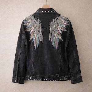 May include: A black denim jacket with a decorative design on the back. The design features colorful embroidered wings with silver fringe. The jacket has a button-down front, two chest pockets, and two side pockets. The collar and cuffs are studded with small silver rivets.