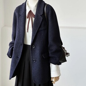 May include: A navy blue blazer with a white collared shirt and a black pleated skirt. The blazer has two buttons and a classic lapel. A burgundy bow tie with white polka dots adds a pop of color. A black shoulder bag is visible.