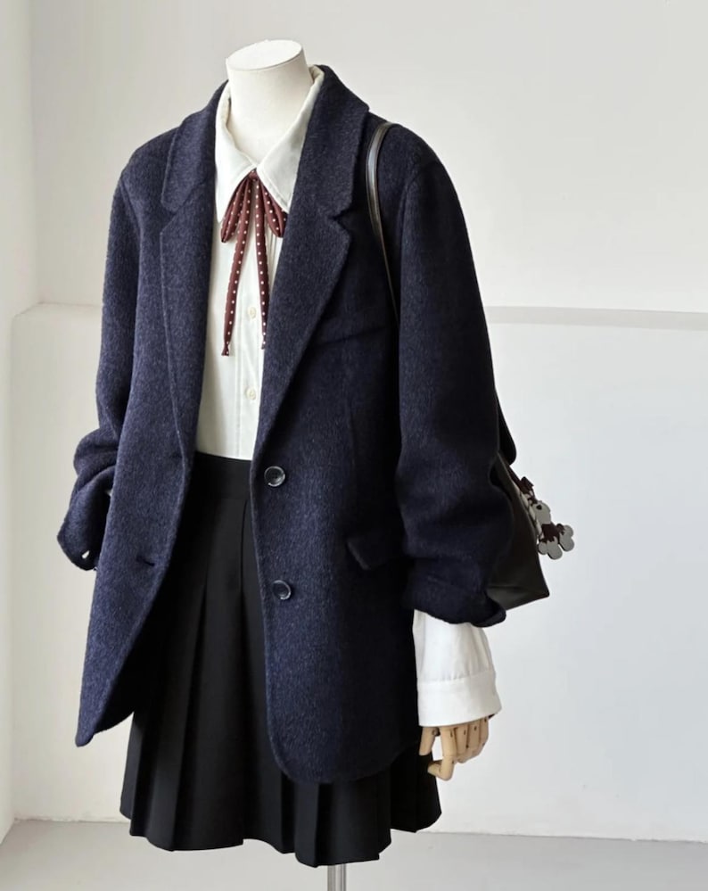May include: A navy blue blazer with a white collared shirt and a black pleated skirt. The blazer has two buttons and a classic lapel. A burgundy bow tie with white polka dots adds a pop of color. A black shoulder bag is visible.