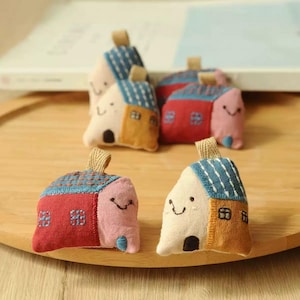 Può includere: Una collezione di ornamenti per la casa in tessuto fatti a mano. Ogni casa presenta un design unico con un volto sorridente, un tetto strutturato e piccoli dettagli delle finestre. Le case sono nei toni del rosso, rosa, blu e beige.