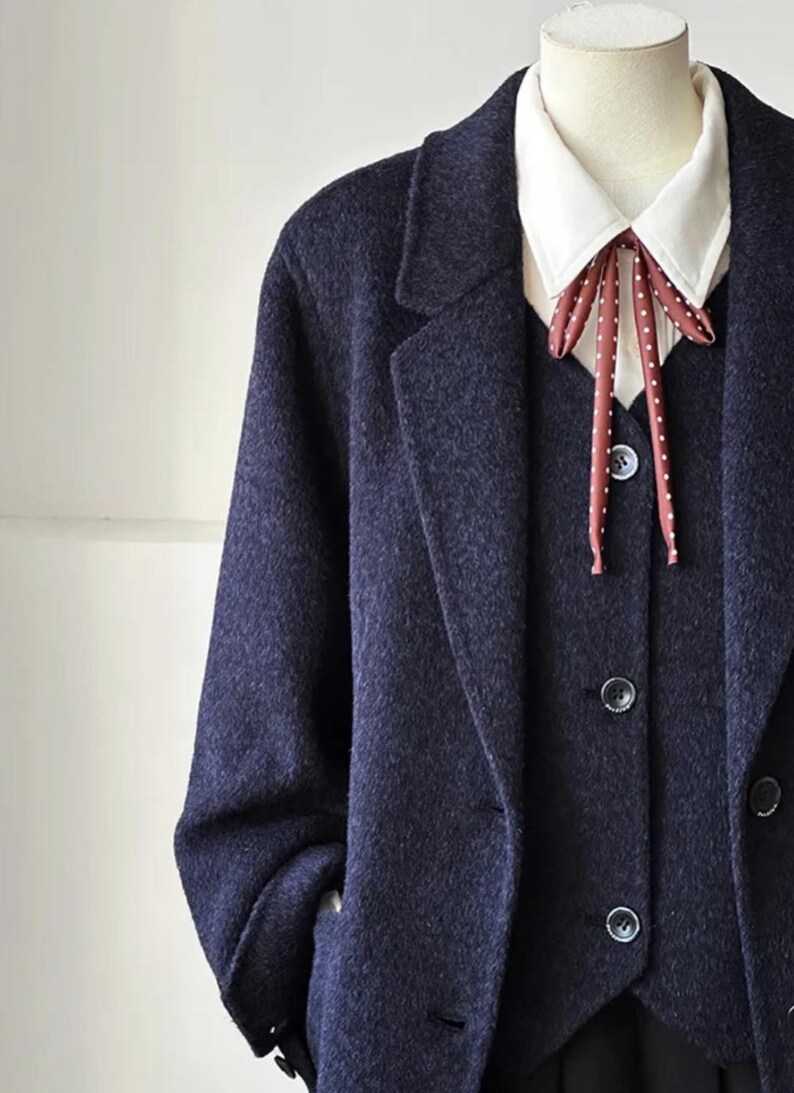 May include: A navy blue suit on a mannequin. The suit includes a long coat, vest, and a white collared shirt with a red and white polka dot bow tie. The suit has a classic, tailored design.