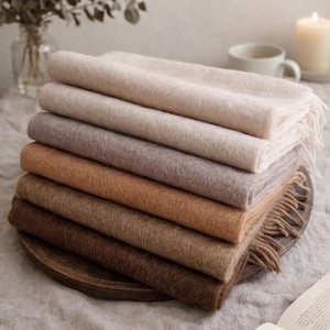 May include: A stack of eight folded scarves in varying shades of beige, brown, and tan. The scarves are arranged on a wooden tray, with the top scarf a light beige and the bottom a deep brown. The edges of the scarves have a fringed detail.