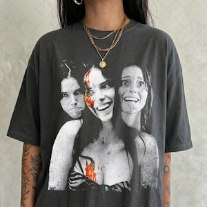 May include: A charcoal gray short-sleeve t-shirt featuring a black and white graphic print of three faces with red accents. The central face is smiling, while the two flanking faces appear distressed. The shirt has a crew neckline and a loose fit.