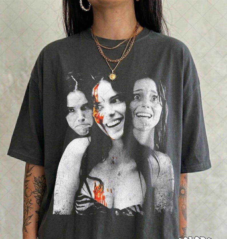 May include: A charcoal gray short-sleeve t-shirt featuring a black and white graphic print of three faces with red accents. The central face is smiling, while the two flanking faces appear distressed. The shirt has a crew neckline and a loose fit.