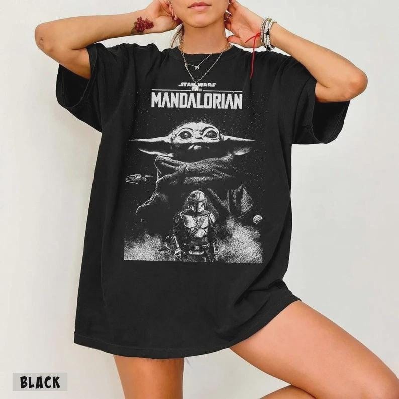 Op de afbeelding: Zwart T-shirt met een afbeelding uit de Star Wars-serie, The Mandalorian. Het ontwerp bevat de tekst "STAR WARS MANDALORIAN" boven een afbeelding van Grogu en de Mandalorian. Het woord "BLACK" staat onderaan.