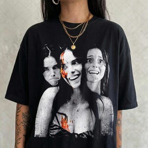 May include: A black short-sleeve t-shirt featuring a large, high-contrast graphic print of three faces with varying expressions. The central face has a splash of red color, and the overall aesthetic is reminiscent of a vintage horror movie poster.