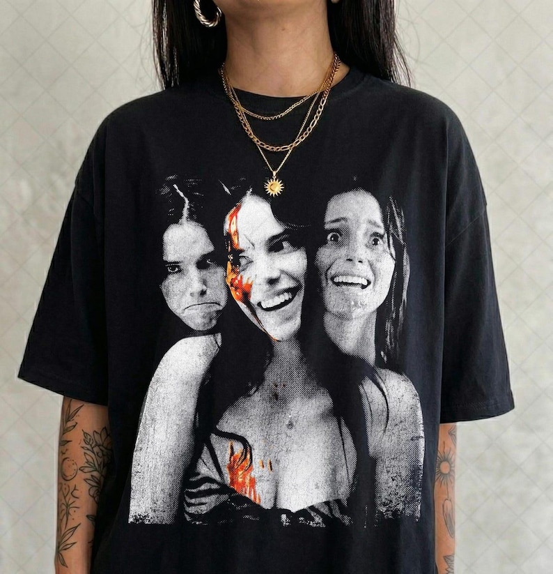 May include: A black short-sleeve t-shirt featuring a large, high-contrast graphic print of three faces with varying expressions. The central face has a splash of red color, and the overall aesthetic is reminiscent of a vintage horror movie poster.