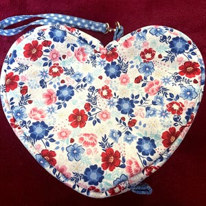 Puede incluir: Bolsa de tela en forma de coraz&oacute;n con un estampado floral en rojo, azul y rosa sobre fondo blanco. La bolsa tiene una correa azul con estrellas blancas y un peque&ntilde;o lazo en la parte inferior. Para llevar objetos peque&ntilde;os.