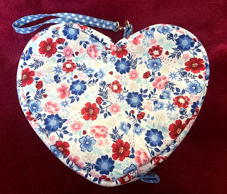 Puede incluir: Bolsa de tela en forma de coraz&oacute;n con un estampado floral en rojo, azul y rosa sobre fondo blanco. La bolsa tiene una correa azul con estrellas blancas y un peque&ntilde;o lazo en la parte inferior. Para llevar objetos peque&ntilde;os.