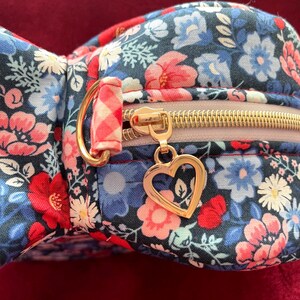 Puede incluir: Un estuche con estampado floral con cremallera dorada y un dije en forma de coraz&oacute;n. El estuche presenta un patr&oacute;n de flores rojas, azules y blancas sobre un fondo oscuro. La cremallera es dorada y el dije es un coraz&oacute;n dorado.
