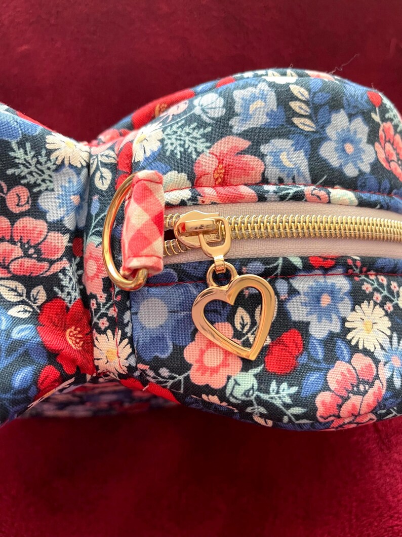 Puede incluir: Un estuche con estampado floral con cremallera dorada y un dije en forma de coraz&oacute;n. El estuche presenta un patr&oacute;n de flores rojas, azules y blancas sobre un fondo oscuro. La cremallera es dorada y el dije es un coraz&oacute;n dorado.