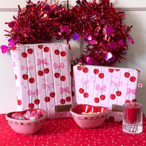May include: Pink quilted pouches with a cherry and bow pattern, displayed with heart-shaped bowls and a red candle. A red tinsel heart-shaped wreath hangs in the background. The pouches have a zipper closure and a small tag that says "RUDY & GOLD".