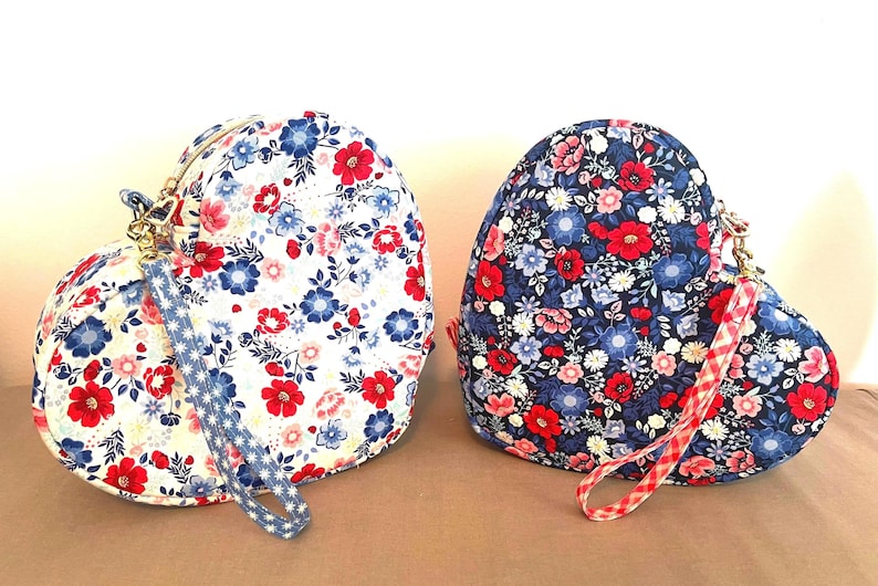 Puede incluir: Dos bolsos de tela con forma de coraz&oacute;n con estampados florales. Uno es blanco con flores rojas y azules, el otro es azul marino con flores rojas y rosas. Cada bolso tiene una correa de mu&ntilde;eca y una cremallera dorada.