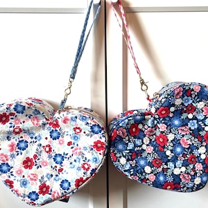 Puede incluir: Dos bolsos en forma de coraz&oacute;n con estampados florales. Uno tiene fondo blanco con flores rojas y azules, el otro fondo azul marino con flores rojas y rosas. Cada bolso tiene una correa a juego y cremallera dorada.