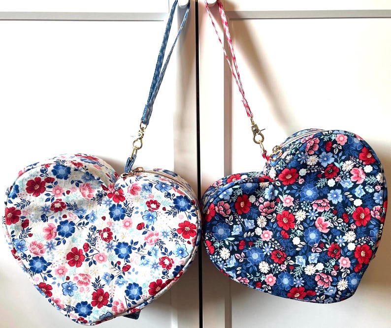 Puede incluir: Dos bolsos en forma de coraz&oacute;n con estampados florales. Uno tiene fondo blanco con flores rojas y azules, el otro fondo azul marino con flores rojas y rosas. Cada bolso tiene una correa a juego y cremallera dorada.