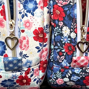 Puede incluir: Primer plano de dos bolsas de tela floral. Una presenta un fondo blanco con flores rojas, rosas y azules, mientras que la otra tiene un fondo azul marino con patrones florales similares. Ambas tienen cremalleras doradas con tiradores en forma de coraz&oacute;n.