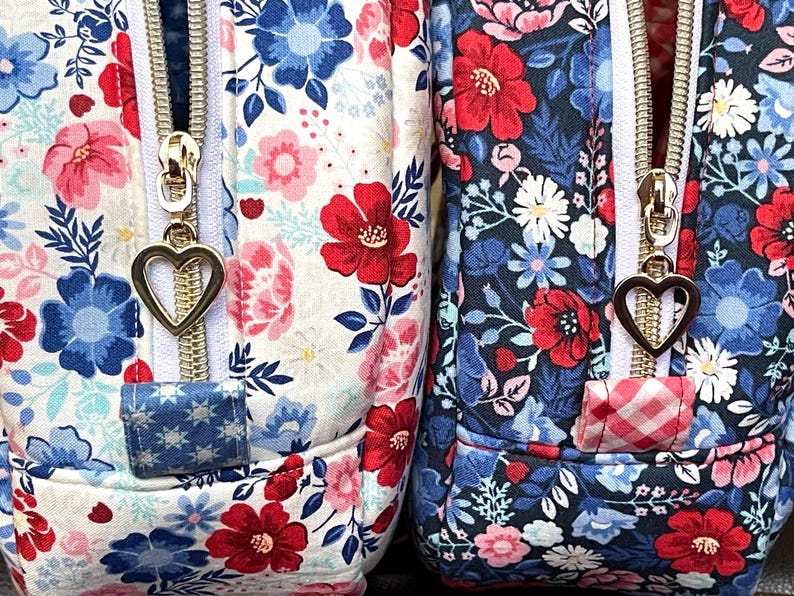 Puede incluir: Primer plano de dos bolsas de tela floral. Una presenta un fondo blanco con flores rojas, rosas y azules, mientras que la otra tiene un fondo azul marino con patrones florales similares. Ambas tienen cremalleras doradas con tiradores en forma de coraz&oacute;n.