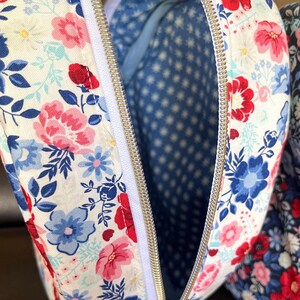 Puede incluir: Primer plano de un neceser con estampado floral y cremallera blanca. El exterior de la bolsa presenta un patr&oacute;n de flores rojas, rosas y azules sobre un fondo blanco. El interior es azul con un patr&oacute;n de estrellas blancas.