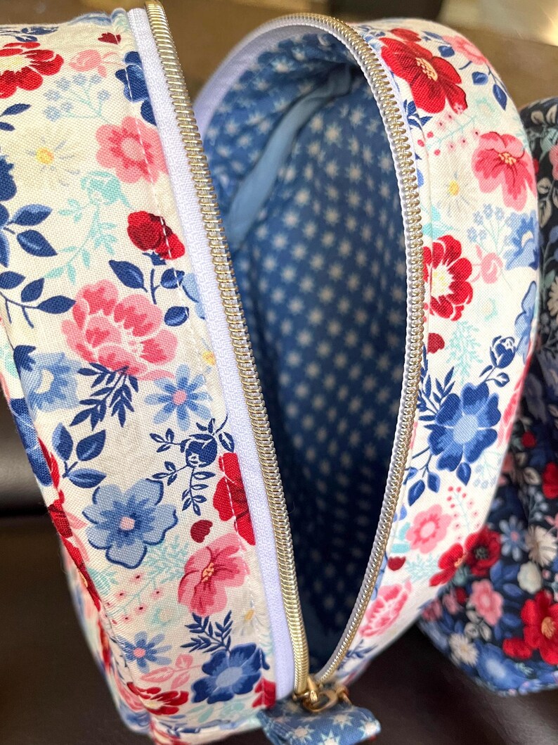 Puede incluir: Primer plano de un neceser con estampado floral y cremallera blanca. El exterior de la bolsa presenta un patr&oacute;n de flores rojas, rosas y azules sobre un fondo blanco. El interior es azul con un patr&oacute;n de estrellas blancas.