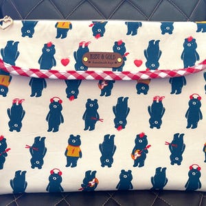 May include: A cream-colored laptop sleeve with a repeating pattern of navy blue bears in various poses. The sleeve has a red and white gingham trim and a brown leather tag that reads "RUDY & GOLD". A gold heart-shaped zipper pull is visible.