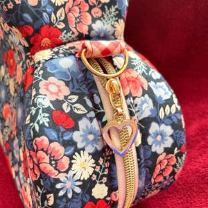 Puede incluir: Un neceser con estampado floral con cremallera dorada y un charm en forma de coraz&oacute;n. La bolsa presenta un estampado de flores rojas, rosas y azules sobre un fondo azul marino. El asa es un anillo dorado con tela a cuadros rojos y blancos.