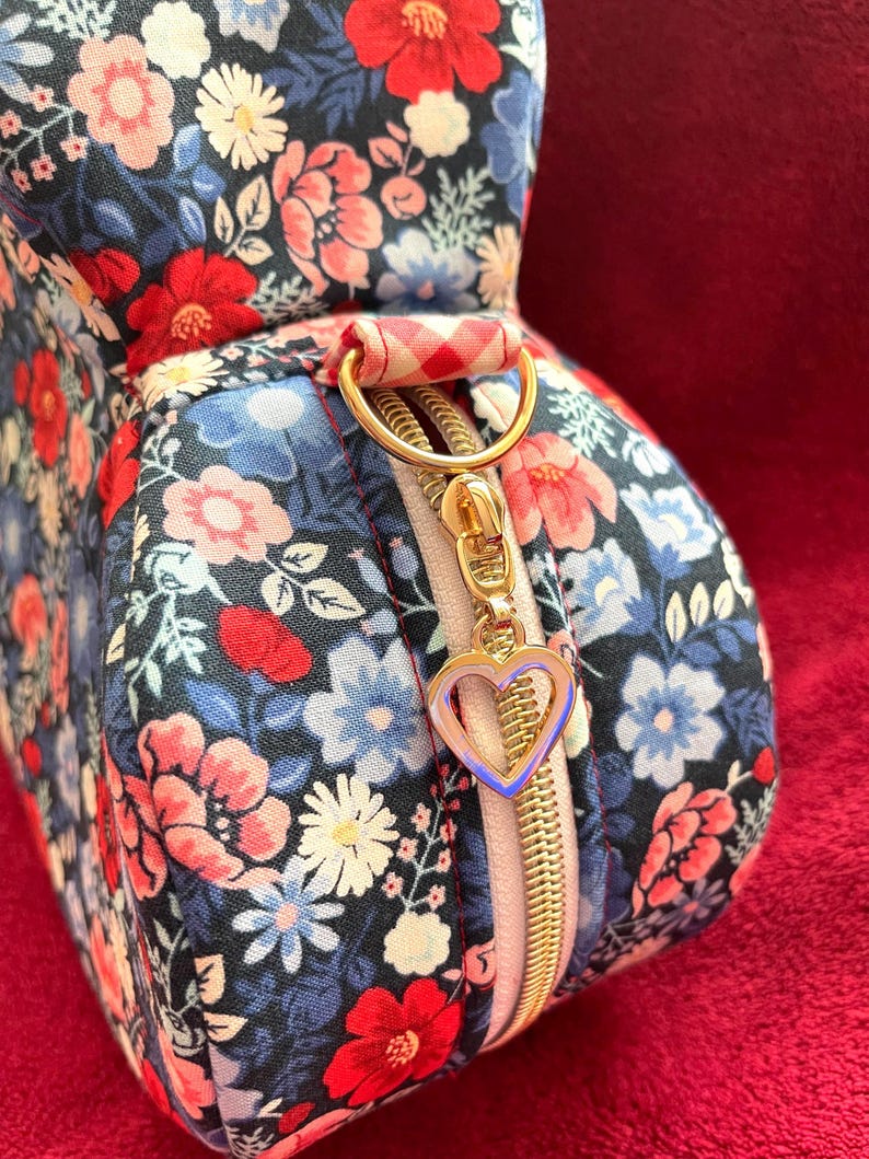 Puede incluir: Un neceser con estampado floral con cremallera dorada y un charm en forma de coraz&oacute;n. La bolsa presenta un estampado de flores rojas, rosas y azules sobre un fondo azul marino. El asa es un anillo dorado con tela a cuadros rojos y blancos.