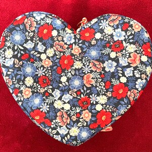Puede incluir: Estuche para accesorios con estampado floral en forma de coraz&oacute;n. El estuche tiene un fondo oscuro con flores rojas, azules y blancas. Se ve una cremallera dorada.