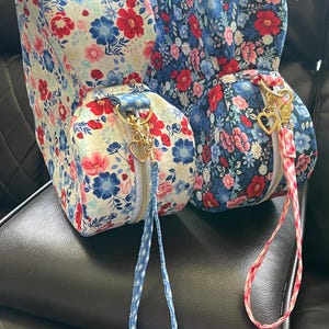 Puede incluir: Dos bolsas con estampado floral, cremalleras doradas y tiradores en forma de coraz&oacute;n. Una bolsa tiene un fondo blanco con flores rojas y azules, la otra un fondo azul con flores rojas y rosas. Ambas tienen correas de mu&ntilde;eca a juego.