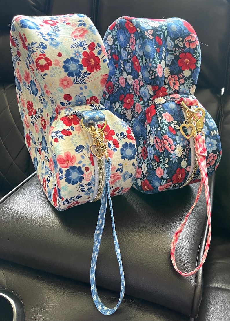 Puede incluir: Dos bolsas con estampado floral, cremalleras doradas y tiradores en forma de coraz&oacute;n. Una bolsa tiene un fondo blanco con flores rojas y azules, la otra un fondo azul con flores rojas y rosas. Ambas tienen correas de mu&ntilde;eca a juego.