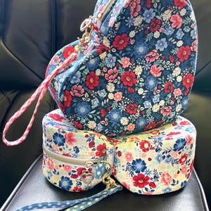 Puede incluir: Tres bolsos con estampado floral apilados. El bolso superior tiene forma de coraz&oacute;n, el del medio es redondo y el inferior tambi&eacute;n. Los bolsos tienen un estampado floral azul marino, rojo y rosa. Cada bolso tiene cremallera y correa de mu&ntilde;eca.