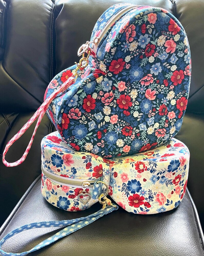 Puede incluir: Tres bolsos con estampado floral apilados. El bolso superior tiene forma de coraz&oacute;n, el del medio es redondo y el inferior tambi&eacute;n. Los bolsos tienen un estampado floral azul marino, rojo y rosa. Cada bolso tiene cremallera y correa de mu&ntilde;eca.