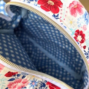 Puede incluir: Vista de cerca de un bolso con estampado floral y cremallera dorada. El exterior del bolso presenta un fondo blanco con flores rojas, rosas y azules. El forro interior es azul con un estampado de estrellas y puntos blancos.