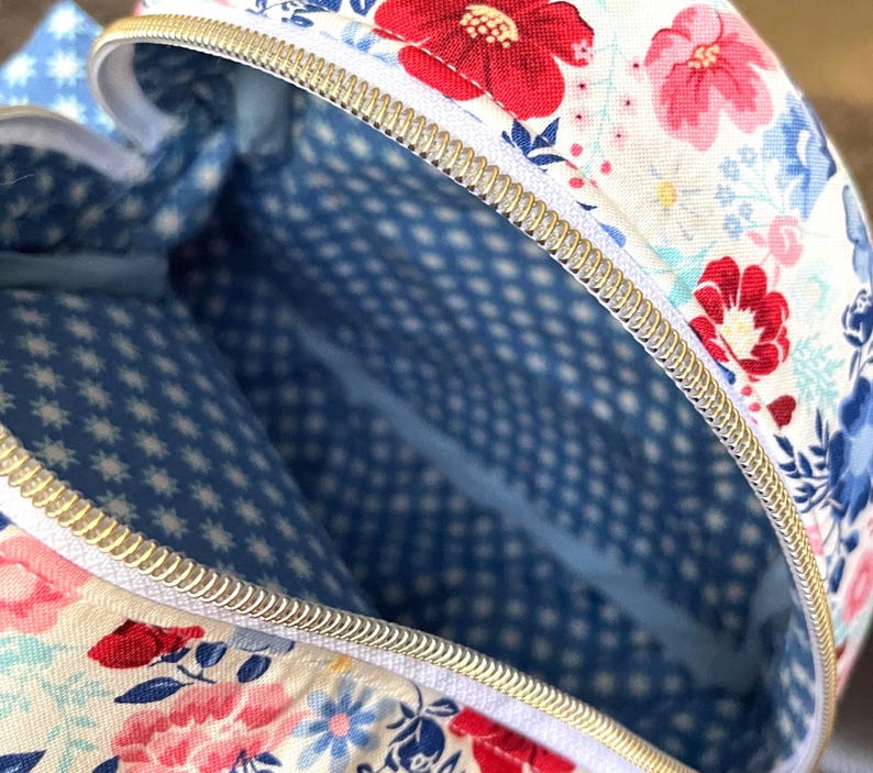 Puede incluir: Vista de cerca de un bolso con estampado floral y cremallera dorada. El exterior del bolso presenta un fondo blanco con flores rojas, rosas y azules. El forro interior es azul con un estampado de estrellas y puntos blancos.