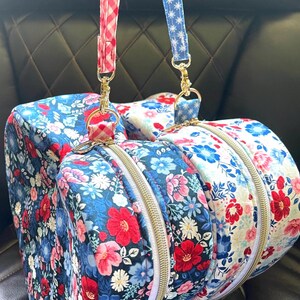 Puede incluir: Dos bolsas de deporte con estampado floral, cremalleras doradas y correas a juego. Una bolsa tiene un fondo azul con flores rojas y rosas, la otra un fondo blanco con flores rojas y azules. Las correas son de cuadros rojos y azules con estrellas blancas.