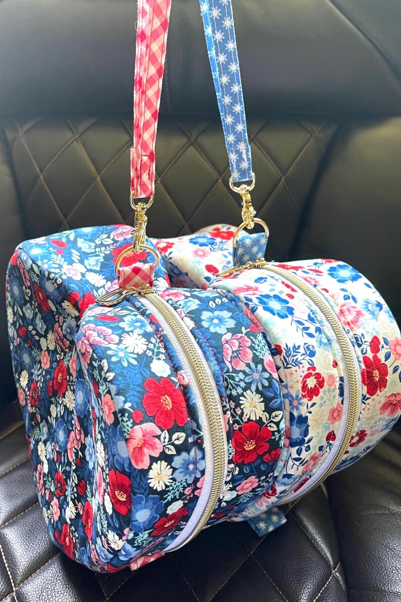 Puede incluir: Dos bolsas de deporte con estampado floral, cremalleras doradas y correas a juego. Una bolsa tiene un fondo azul con flores rojas y rosas, la otra un fondo blanco con flores rojas y azules. Las correas son de cuadros rojos y azules con estrellas blancas.