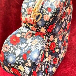 Puede incluir: Un neceser con estampado floral con cremallera dorada y un dije en forma de coraz&oacute;n. La bolsa presenta un patr&oacute;n de flores rojas, azules y blancas sobre un fondo oscuro. La bolsa es rectangular con bordes redondeados.