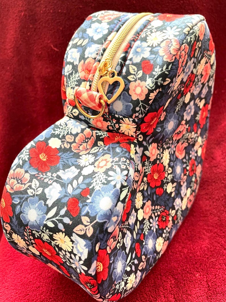 Puede incluir: Un neceser con estampado floral con cremallera dorada y un dije en forma de coraz&oacute;n. La bolsa presenta un patr&oacute;n de flores rojas, azules y blancas sobre un fondo oscuro. La bolsa es rectangular con bordes redondeados.