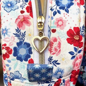 Puede incluir: Primer plano de un neceser con estampado floral. La bolsa presenta un fondo blanco con flores rojas, azules y rosas. Una cremallera dorada con un tirador en forma de coraz&oacute;n est&aacute; centrada. Una peque&ntilde;a leng&uuml;eta de tela azul con motivos de estrellas blancas se encuentra en la base de la cremallera.