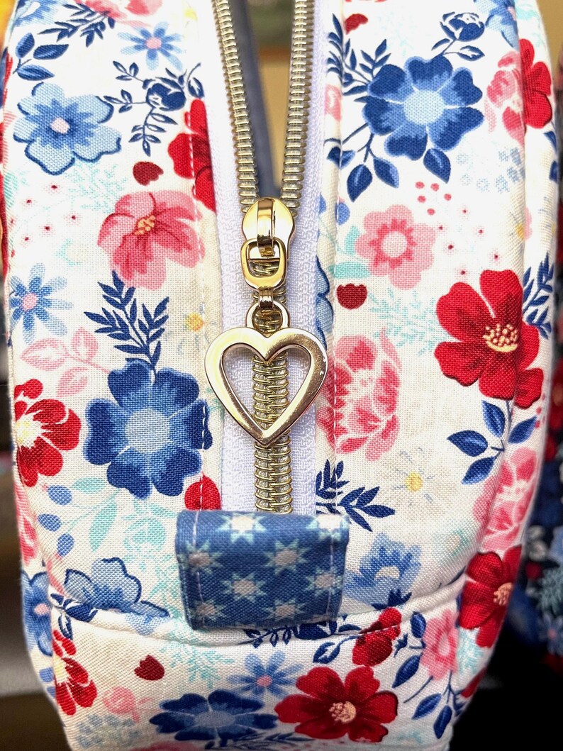 Puede incluir: Primer plano de un neceser con estampado floral. La bolsa presenta un fondo blanco con flores rojas, azules y rosas. Una cremallera dorada con un tirador en forma de coraz&oacute;n est&aacute; centrada. Una peque&ntilde;a leng&uuml;eta de tela azul con motivos de estrellas blancas se encuentra en la base de la cremallera.