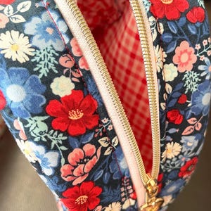 Puede incluir: Bolsa de cosm&eacute;ticos con estampado floral y cremallera dorada. La bolsa tiene un fondo azul marino con flores rojas, azules y blancas. El forro interior es de cuadros rojos y blancos.