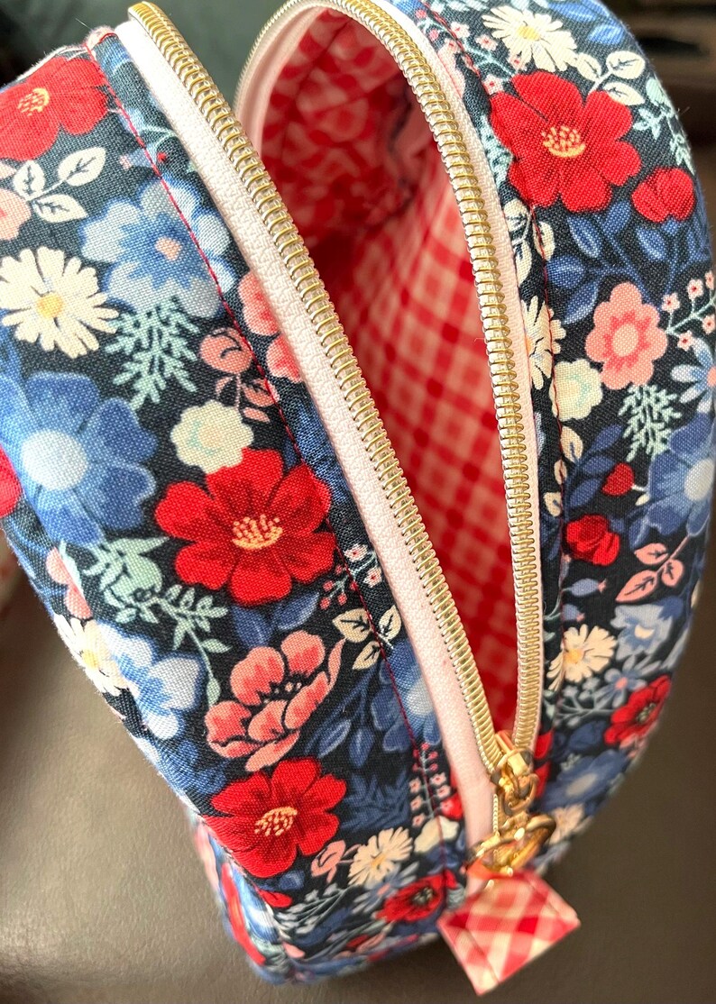 Puede incluir: Bolsa de cosm&eacute;ticos con estampado floral y cremallera dorada. La bolsa tiene un fondo azul marino con flores rojas, azules y blancas. El forro interior es de cuadros rojos y blancos.