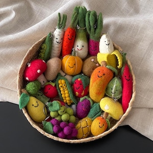 May include: A wicker basket filled with a variety of felted fruit and vegetable keyrings. The collection features carrots, radishes, pumpkins, corn, grapes, and more, each with smiling faces. The keyrings are in shades of red, orange, yellow, green, and purple.