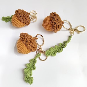 Op de afbeelding: Drie gehaakte eikel-sleutelhangers met lichtbruine garen lichamen en donkerbruine, getextureerde dopjes. Elke eikel is bevestigd aan een goudkleurige metalen ring. Twee van de sleutelhangers bevatten een lang, groen gehaakt eikenblad.