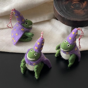 Può includere: Tre ornamenti a forma di rana verde in feltro ad ago, ognuno con un cappello a punta e un mantello viola decorati con stelle gialle ricamate. Ogni rana presenta una pancia bianca e un cordino ritorto rosso e bianco sulla punta del cappello.