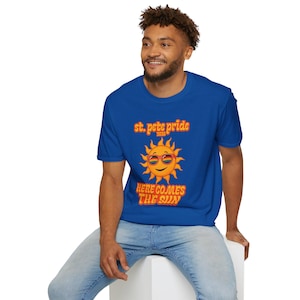 Puede incluir: Camiseta azul real con el texto "st. pete pride 2020" en letras naranjas. Debajo, un sol sonriente con gafas de sol de arcoíris. La frase "HERE COMES THE SUN" está escrita debajo del sol en naranja.