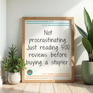 May include: A framed white sign with the text "Not procrastinating. Just reading 500 reviews before buying a stapler." The frame is light brown, and the text is in black. The sign has a teal and orange border. Potted plants flank the sign.